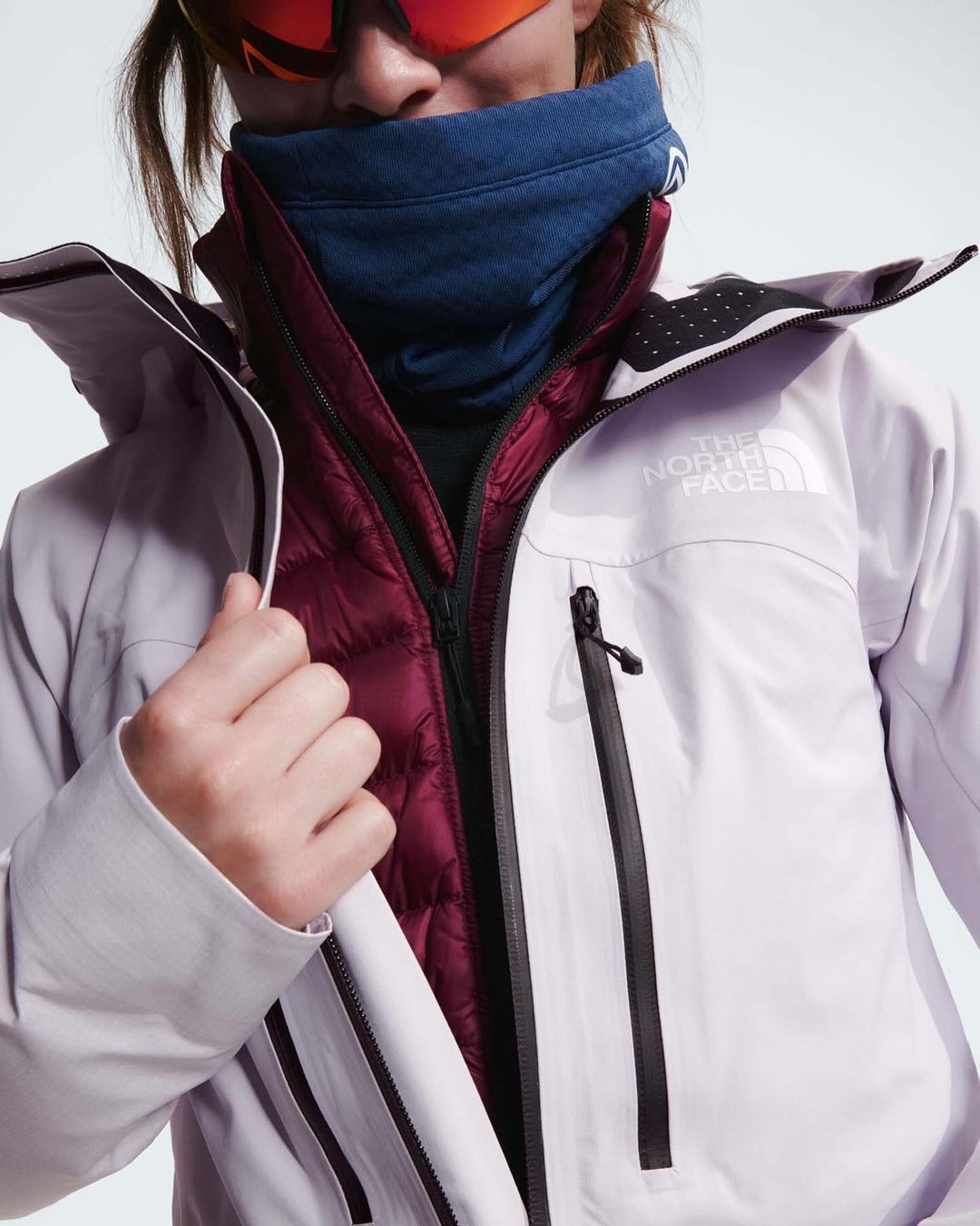 Close-up of a person wearing a light purple North Face jacket with a burgundy quilted inner layer, sunglasses, and blue neck warmer.
