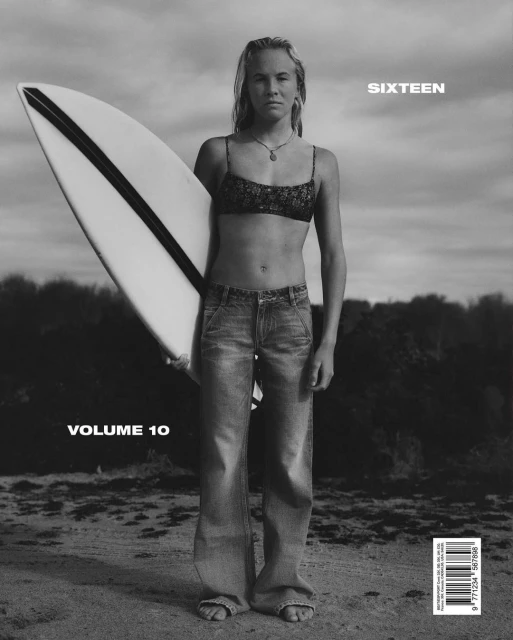 Teen female surfer stands on the beach holding surfboard in black and white, fashion magazine cover with 'SIXTEEN' and 'VOLUME 10' visible.