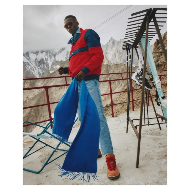 Man in red jacket, jeans, and boots stands near ski rack in a mountain setting, holding a long blue scarf, posing for a fashion shot.