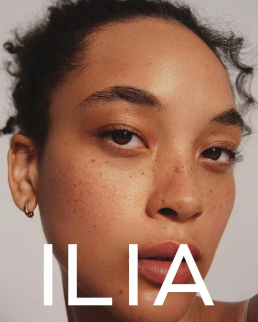 Close-up portrait of a young woman with freckles and curly hair against a white background featuring the 'ILIA' brand logo.