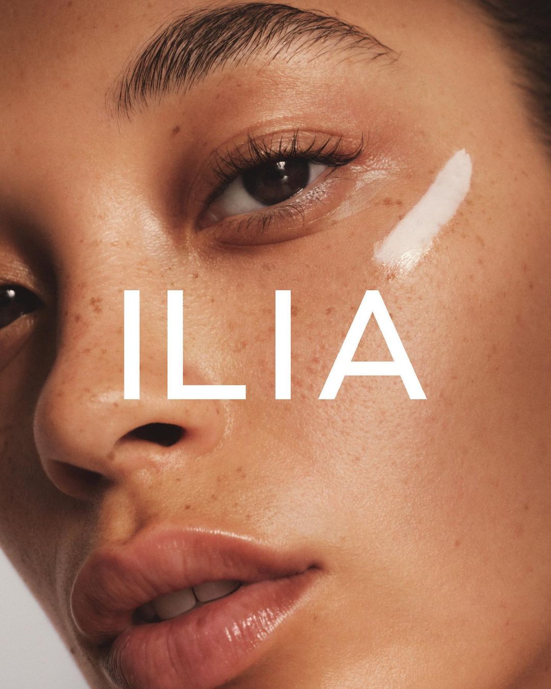 Close-up of an adult woman's face featuring smooth skin, visible freckles, a white cosmetic product, and the brand name ILIA in white letters.
