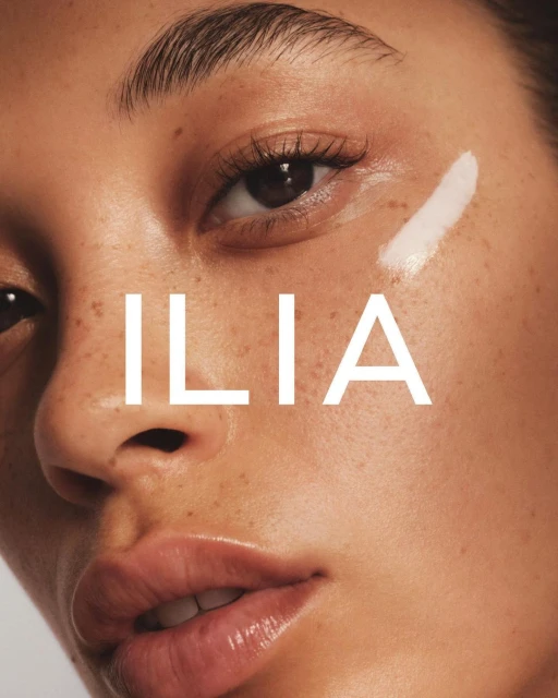 Close-up of an adult woman's face featuring smooth skin, visible freckles, a white cosmetic product, and the brand name ILIA in white letters.