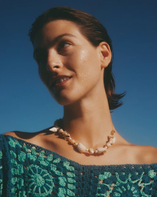 Close-up of a young adult woman with wet hair, wearing a shell necklace and crocheted top against a clear blue sky backdrop.