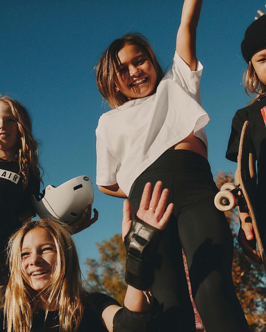 Group of young people smiling and enjoying a day outdoors, with skateboards and protective gear. The clear blue sky provides backdrop.