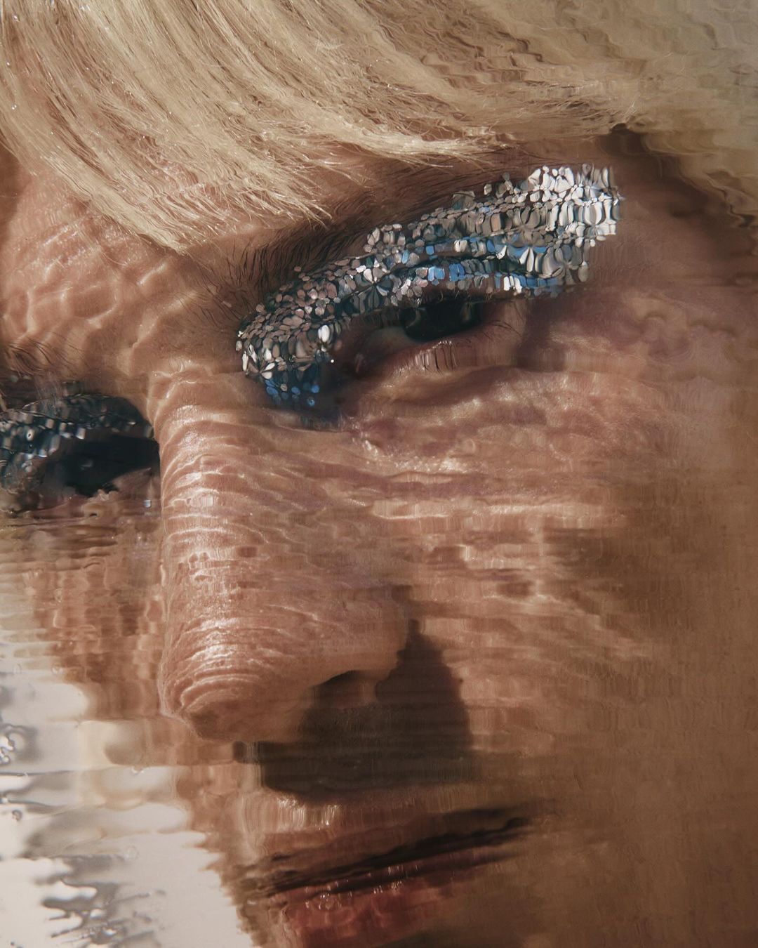 Close-up of woman's face with blond hair and makeup; silver glitter applied around the eyes, with a rippled glass or water effect overlay.