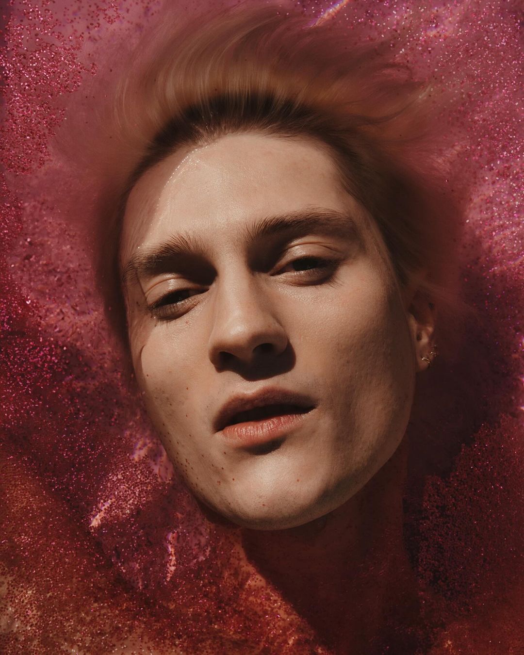 Close-up portrait of a man submerged in shimmering magenta water with floating glitter particles by photographers Ward & Kweskin.