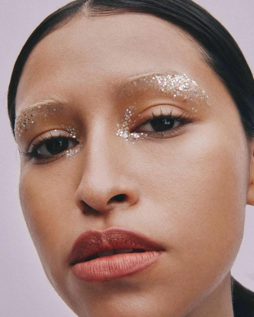 Close-up of a woman with glitter makeup around her eyes and eyebrows against a light purple background in a studio portrait.