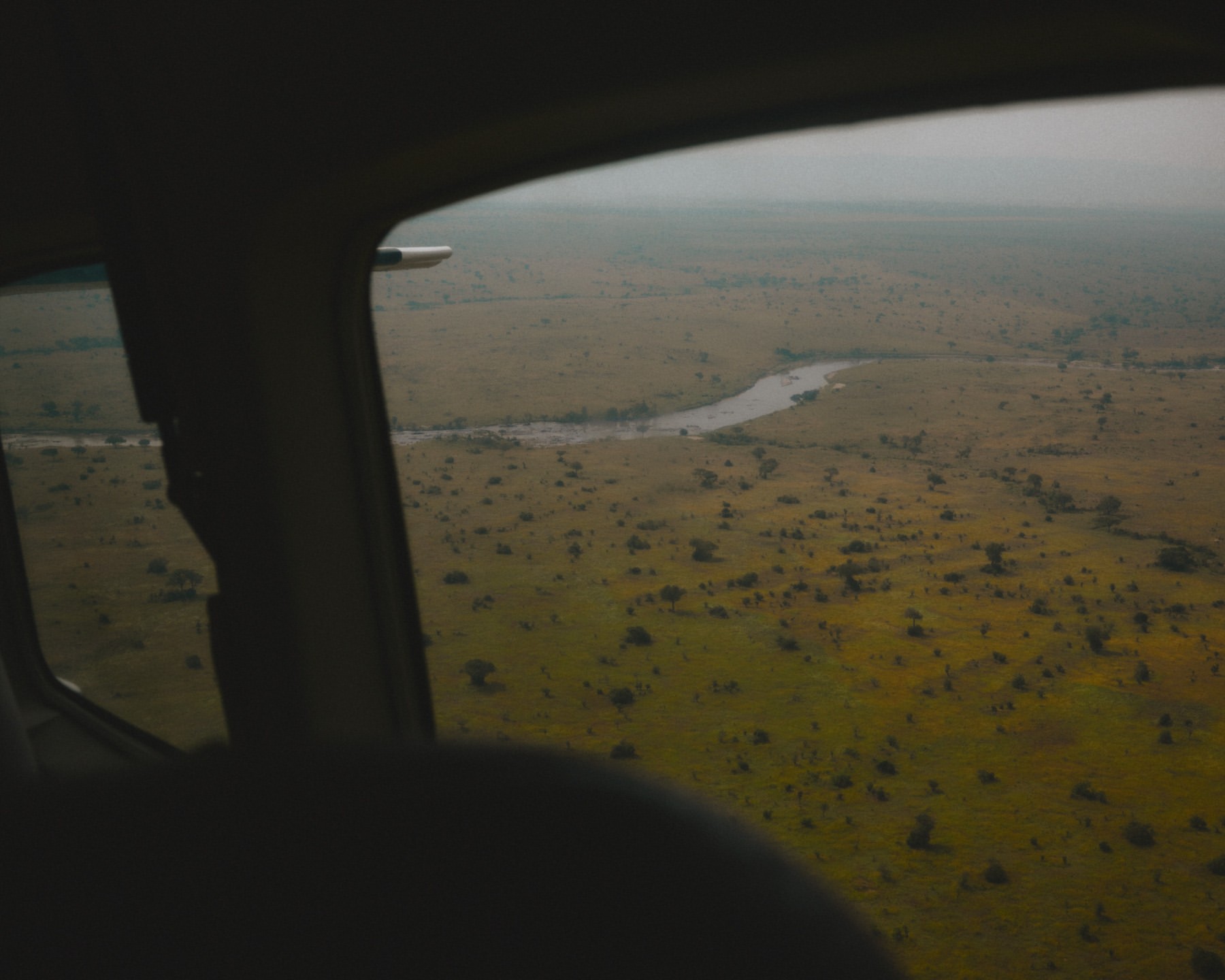Aerial view from inside airplane of river winding through savanna landscape with trees and sky in the background, visible through window.