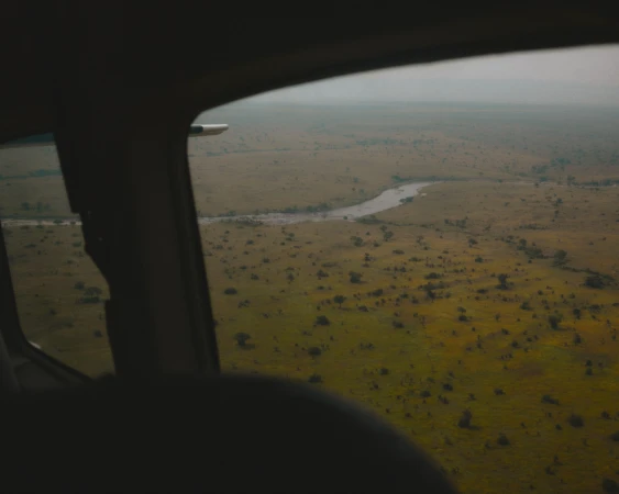 Aerial view from inside airplane of river winding through savanna landscape with trees and sky in the background, visible through window.