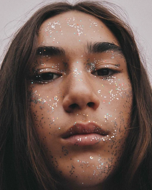 Close-up portrait of a woman with long brown hair and glitter freckles on her face against a bright, neutral background.