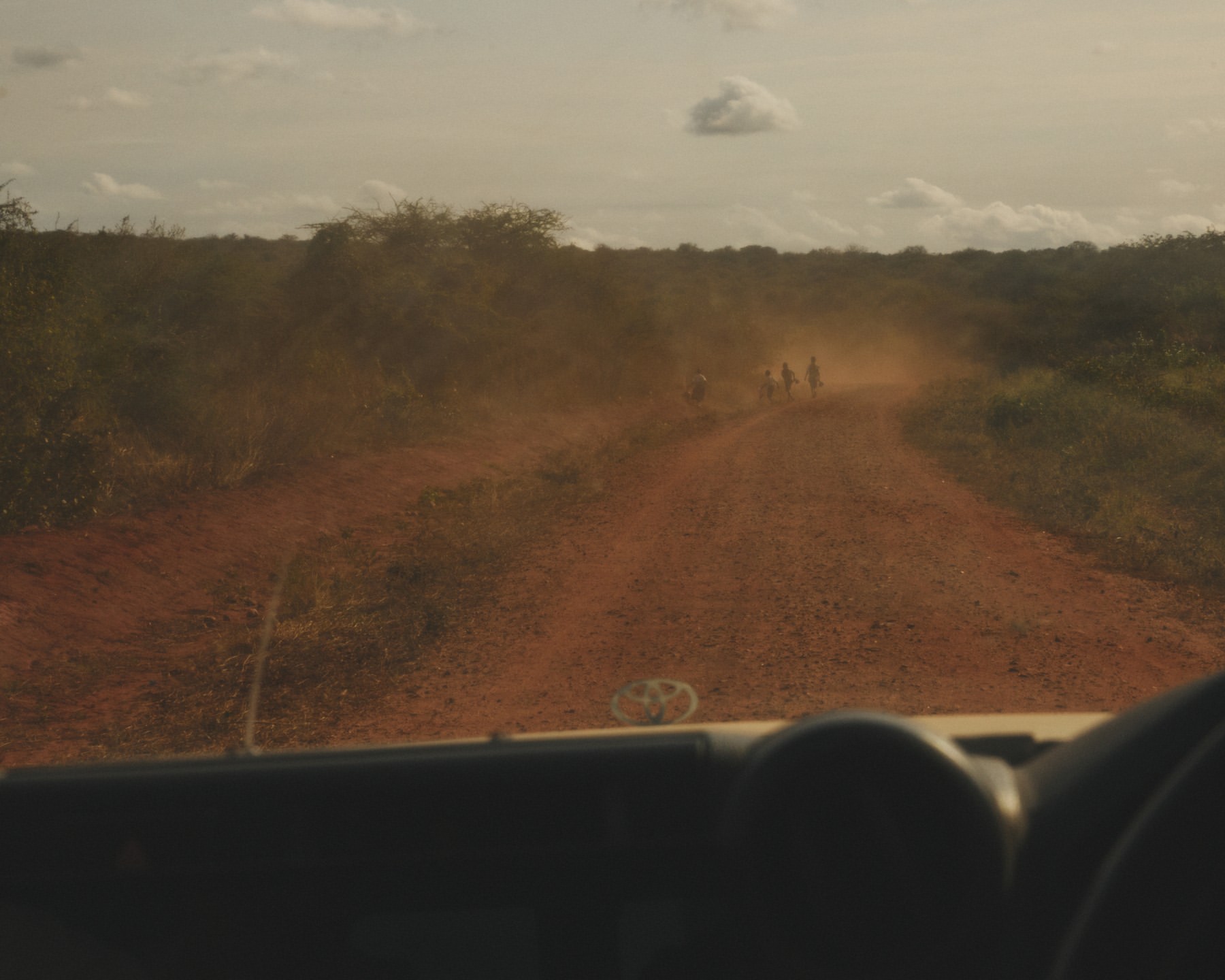 Dusty rural road with group of people in distance, viewed from inside vehicle with dashboard in foreground, and savanna-like vegetation.