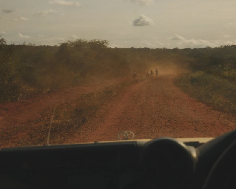 Dusty rural road with group of people in distance, viewed from inside vehicle with dashboard in foreground, and savanna-like vegetation.