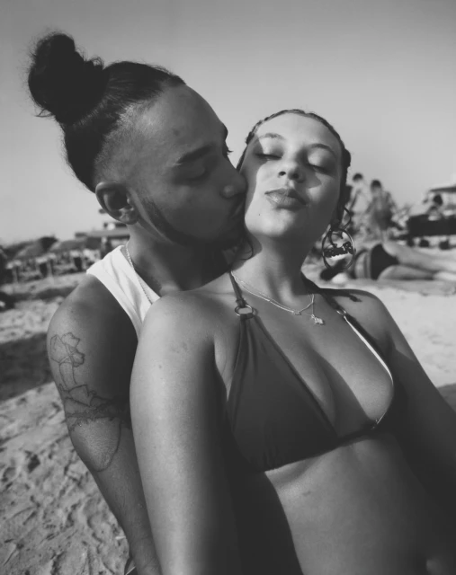 Monochrome image of a couple embracing on a beach, with other people visible in the background. The man is kissing the woman's cheek.