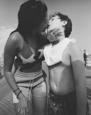 Black and white close-up of two people in swimwear embracing on a wooden boardwalk with buildings and sky in the background.