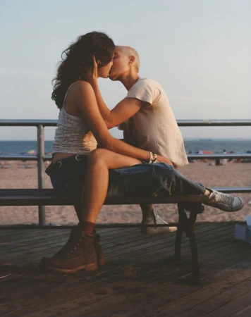 Two people kissing while sitting together on a wooden bench on a boardwalk overlooking the ocean at sunset.