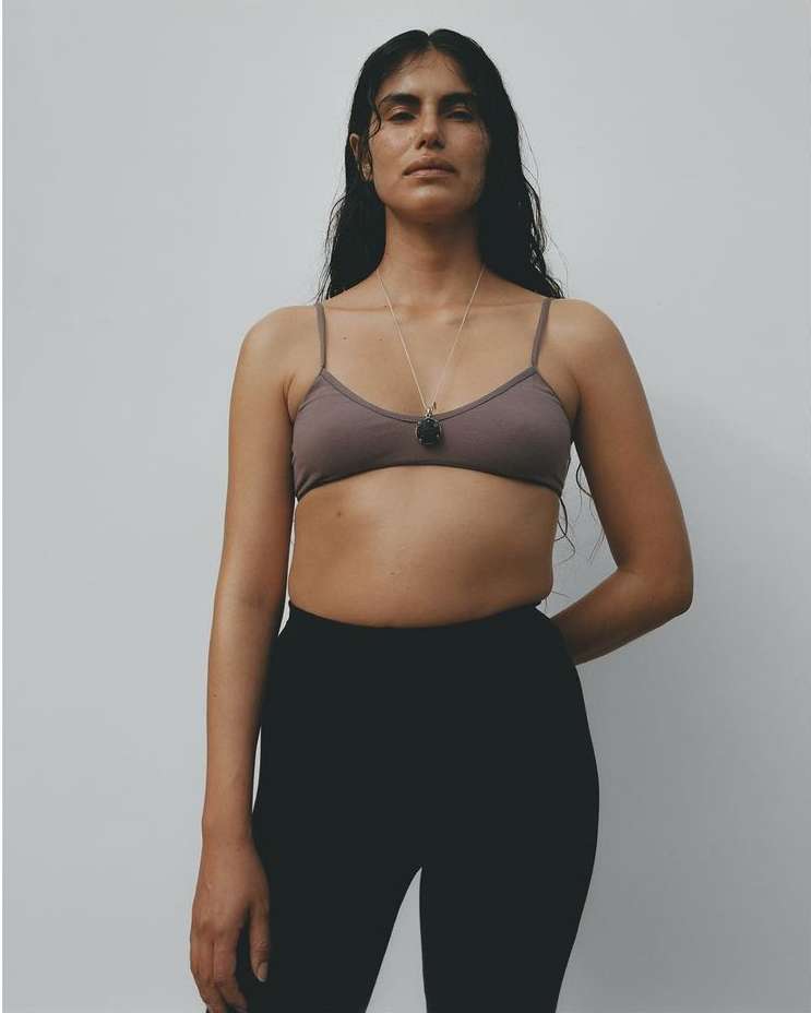 Standing studio portrait of a brunette adult woman wearing a bra top and black leggings, facing forward with a neutral expression.