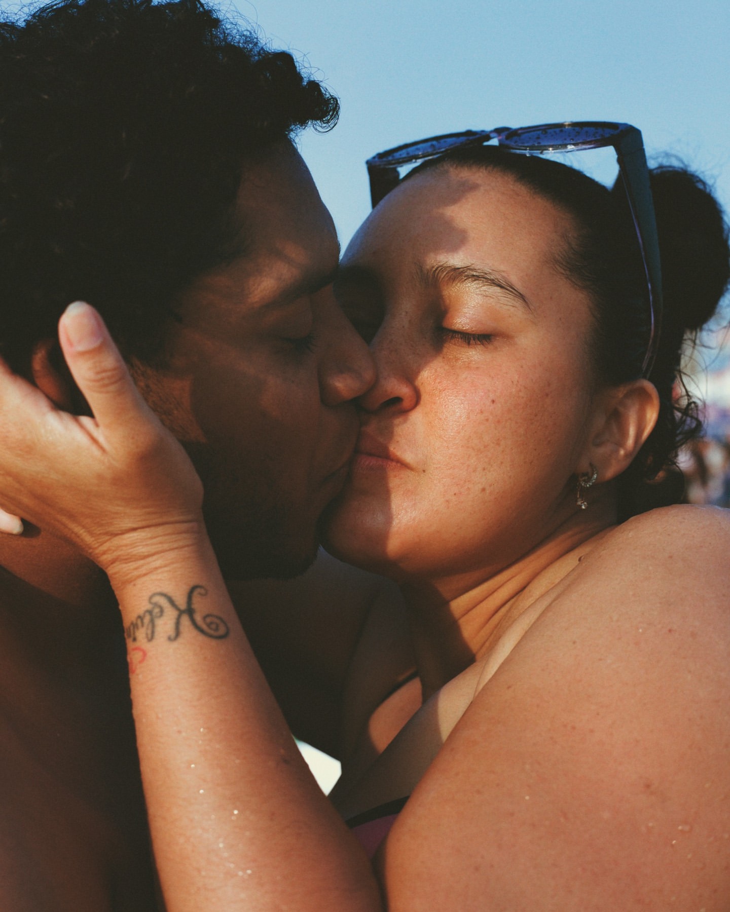 Close-up shot of a romantic couple kissing on a sunny beach, with clear blue sky and a blurry background of beachgoers on a bright, sunny day.