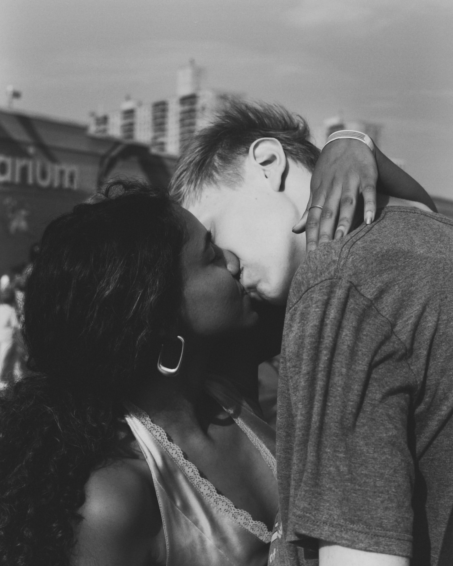 Monochrome portrait of a couple in a close embrace, sharing a kiss in an outdoor setting with buildings in the background.