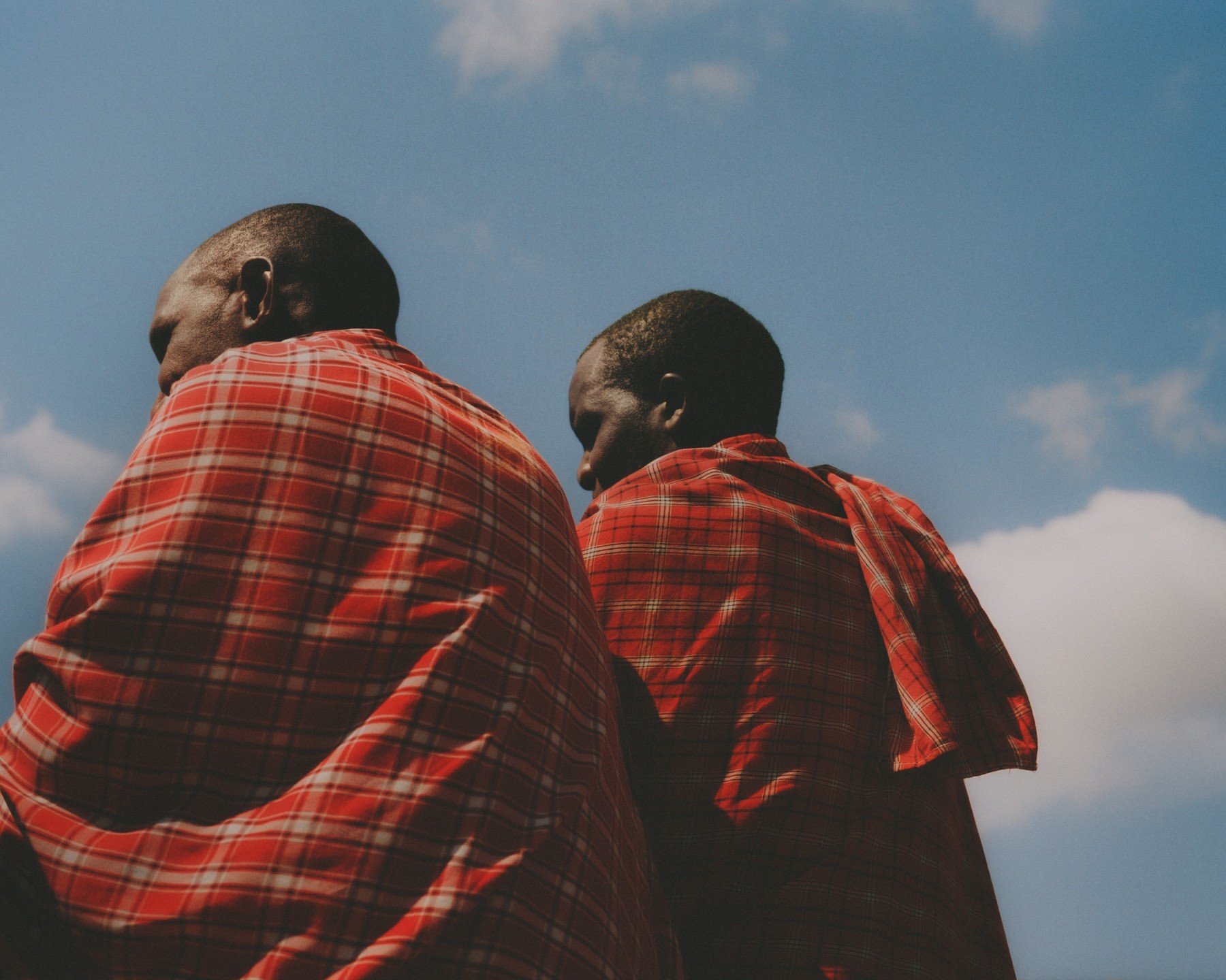 Two adult Masai men in red plaid shuka garments stand outside against a partly cloudy blue sky, low angle, outdoors, daytime.