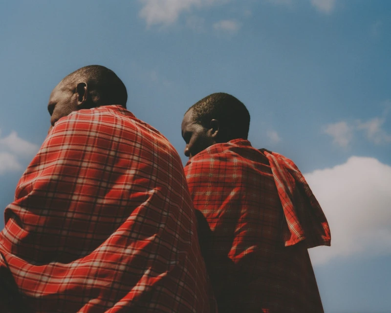 Two adult Masai men in red plaid shuka garments stand outside against a partly cloudy blue sky, low angle, outdoors, daytime.