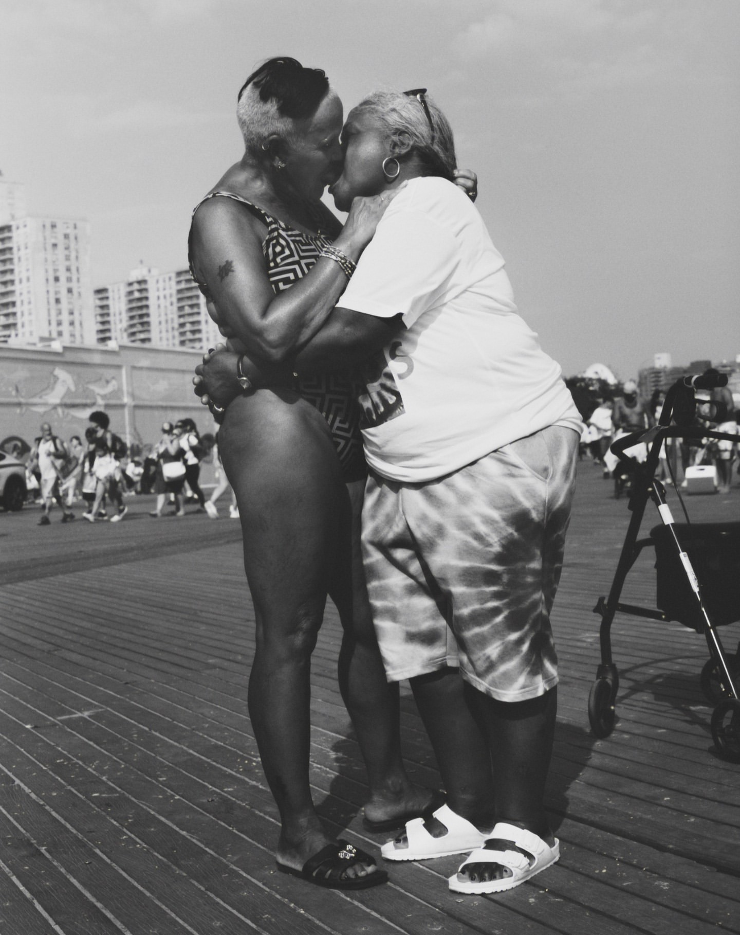 Black and white photograph of two adult women embracing and kissing on an outdoor boardwalk near buildings, showcasing an intimate moment.
