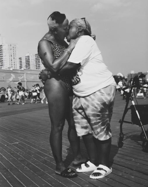 Black and white photograph of two adult women embracing and kissing on an outdoor boardwalk near buildings, showcasing an intimate moment.