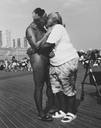 Black and white photograph of two adult women embracing and kissing on an outdoor boardwalk near buildings, showcasing an intimate moment.
