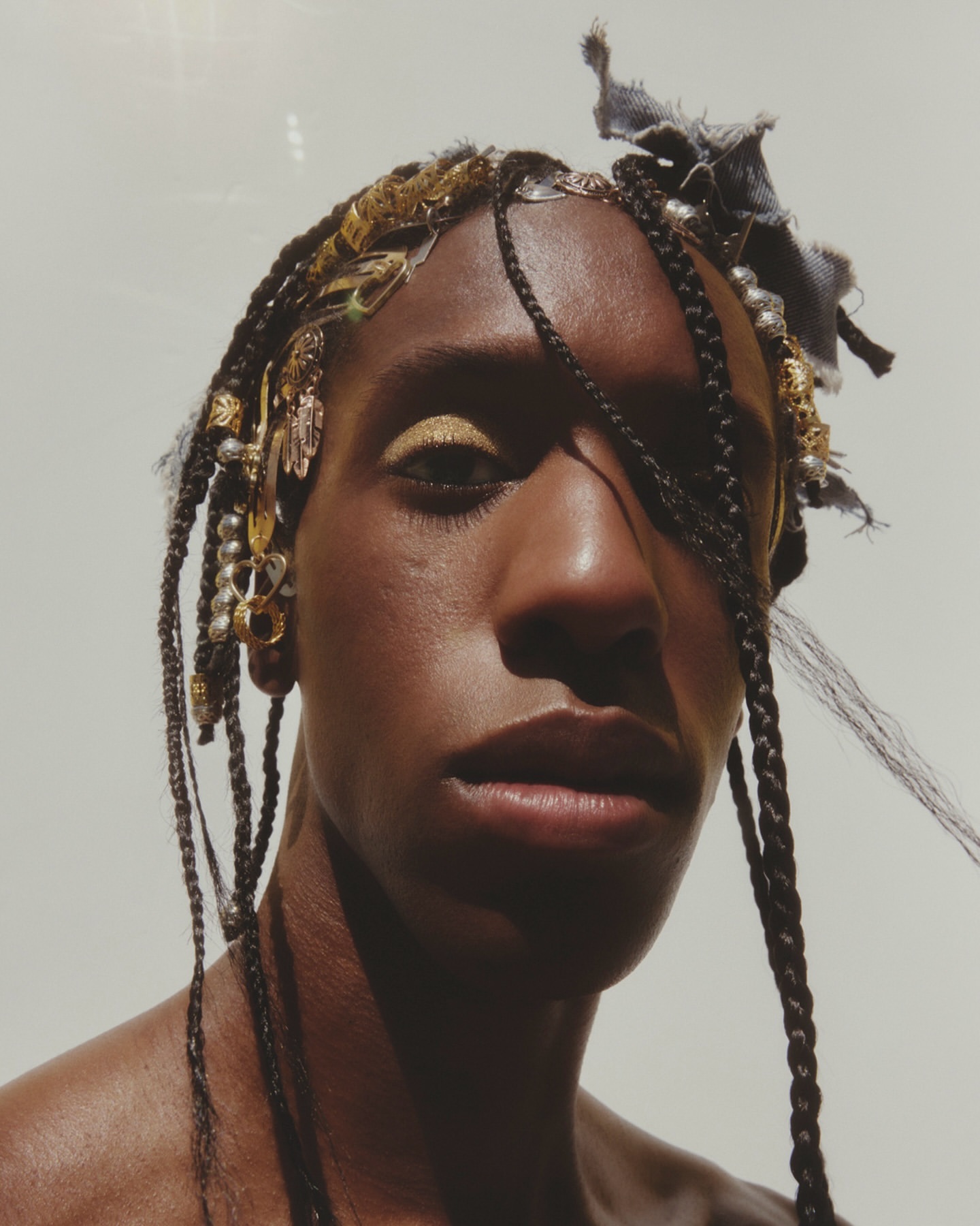 Close-up of an adult with braids, adorned with gold jewelry and fabric, posing indoors with gold eyeshadow against a light background.
