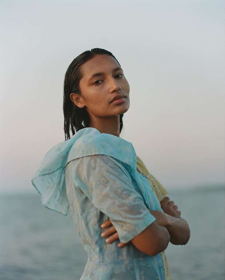 Portrait of a woman with wet hair and arms crossed, wearing a light blue dress, standing in front of a body of water and light sky.