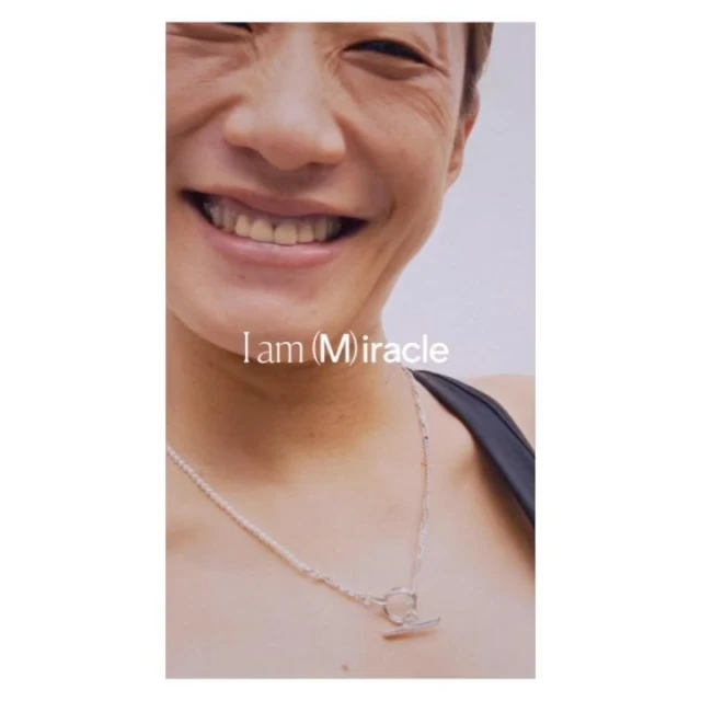 Close-up of a smiling adult wearing a silver chain necklace with a bar pendant; text overlay reads “I am (M)iracle.