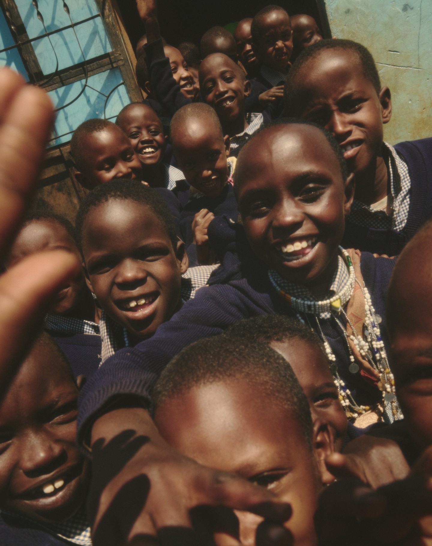 A large group of smiling African children in school uniforms are seen close up, some waving their hands, in an indoor environment.