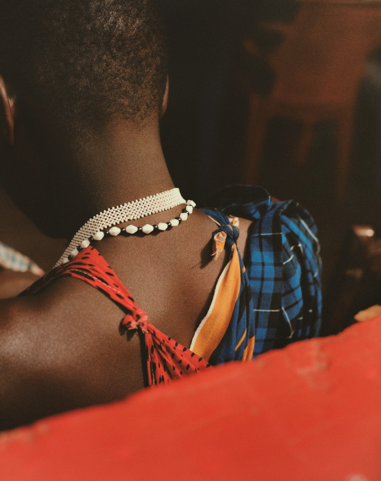 Back view of an African person wearing a blue and orange top and layered necklaces. The focus is on the back and shoulders.