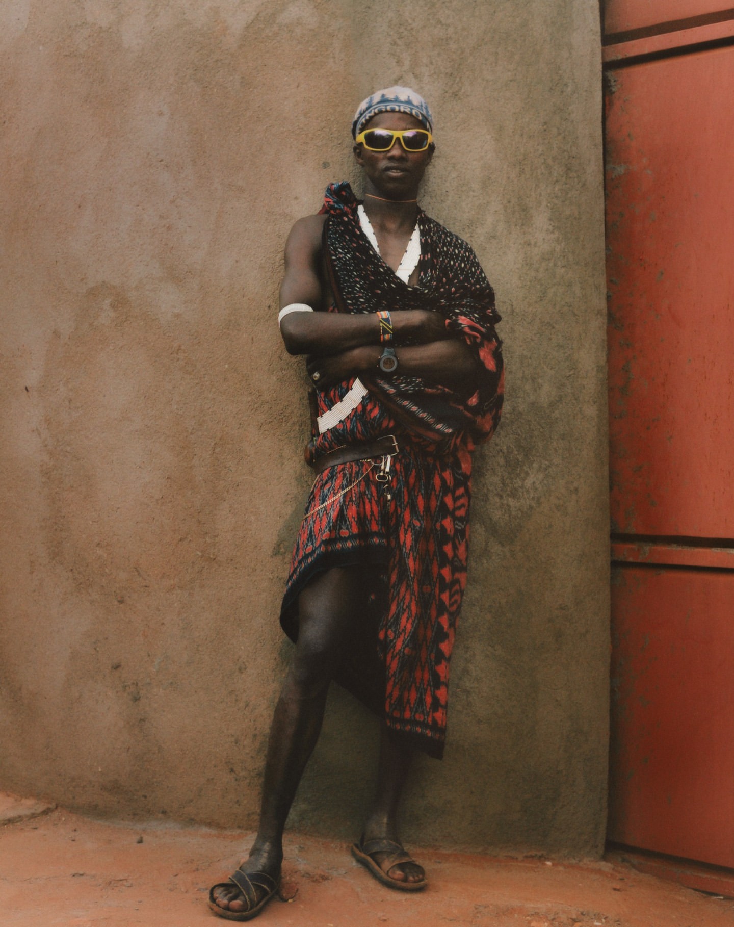 Portrait of an African man with yellow sunglasses and a patterned sarong, standing against a concrete wall with a confident pose.