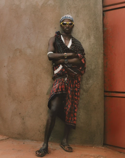 Portrait of an African man with yellow sunglasses and a patterned sarong, standing against a concrete wall with a confident pose.