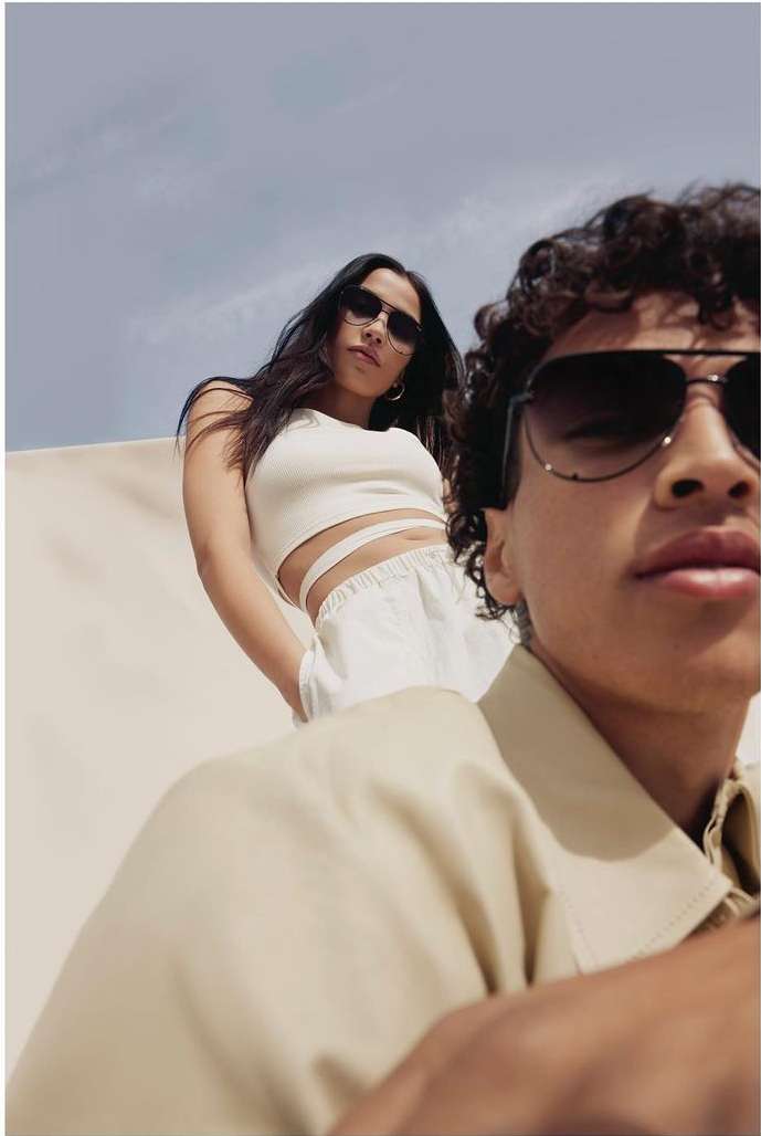 Two young adults model sunglasses outdoors against a neutral backdrop with a clear sky, showcasing a modern fashion aesthetic.