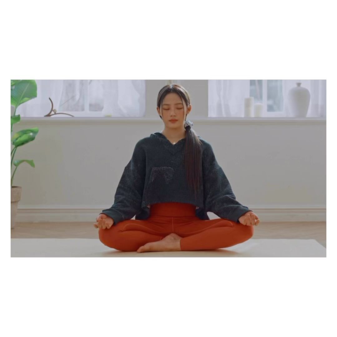 Woman in meditative lotus pose indoors.  She is wearing an athleisure outfit in a bright, neutral space with natural light and green plant.