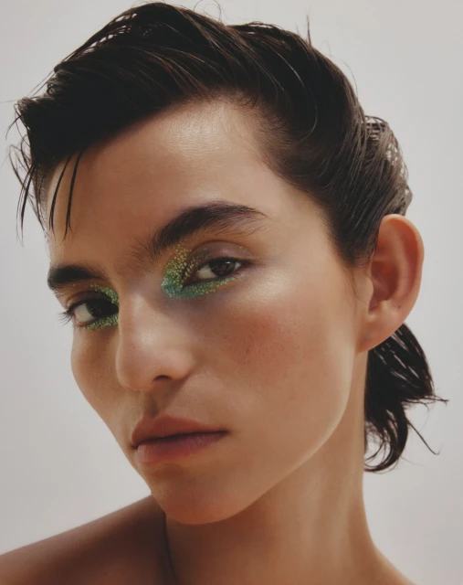 Glamorous close-up of a woman with striking green and gold glitter eye makeup, wet slicked-back hair, and a clean, bright complexion.