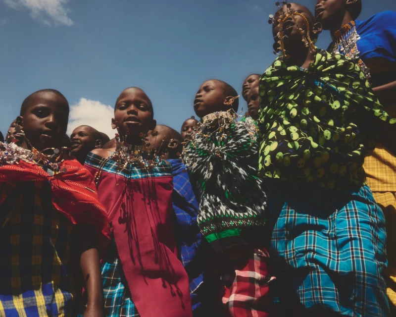 Group of African children in vibrant, traditional garments and beaded necklaces looking directly at the camera against a bright sky.