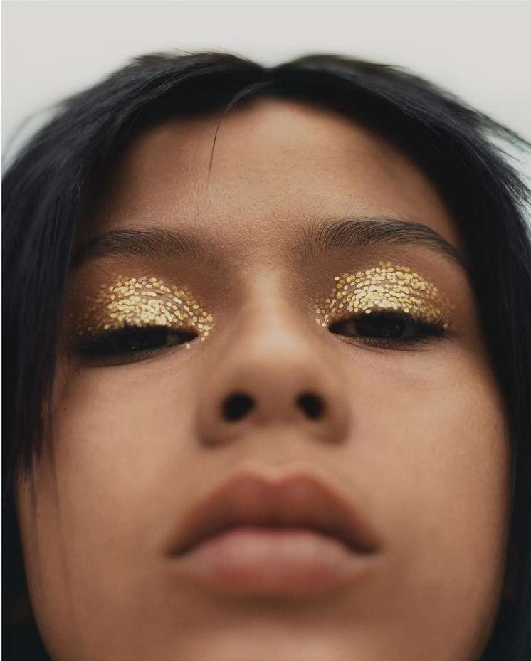 Close-up shot of a young adult woman with dramatic gold glitter eye makeup. Low angle, dark hair, fair skin, neutral facial expression.