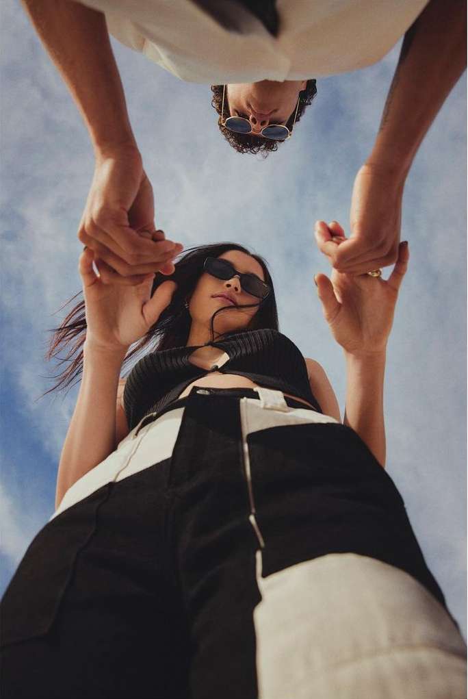 Low-angle shot of a stylish couple holding hands against a vibrant blue sky, featuring bold fashion choices and clear sky backdrop.