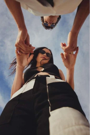 Low-angle shot of a stylish couple holding hands against a vibrant blue sky, featuring bold fashion choices and clear sky backdrop.