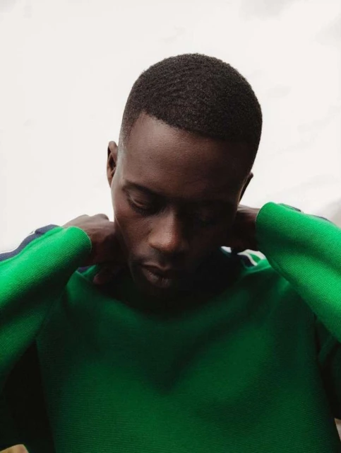 Indoor portrait of a Black man in a green sweater, looking down with hands near his neck. Neutral background, medium shot.