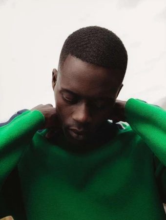 Indoor portrait of a Black man in a green sweater, looking down with hands near his neck. Neutral background, medium shot.