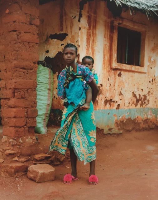 Young African girl carries baby on her back, standing outside a mud brick building with a thatched roof in a rural setting.