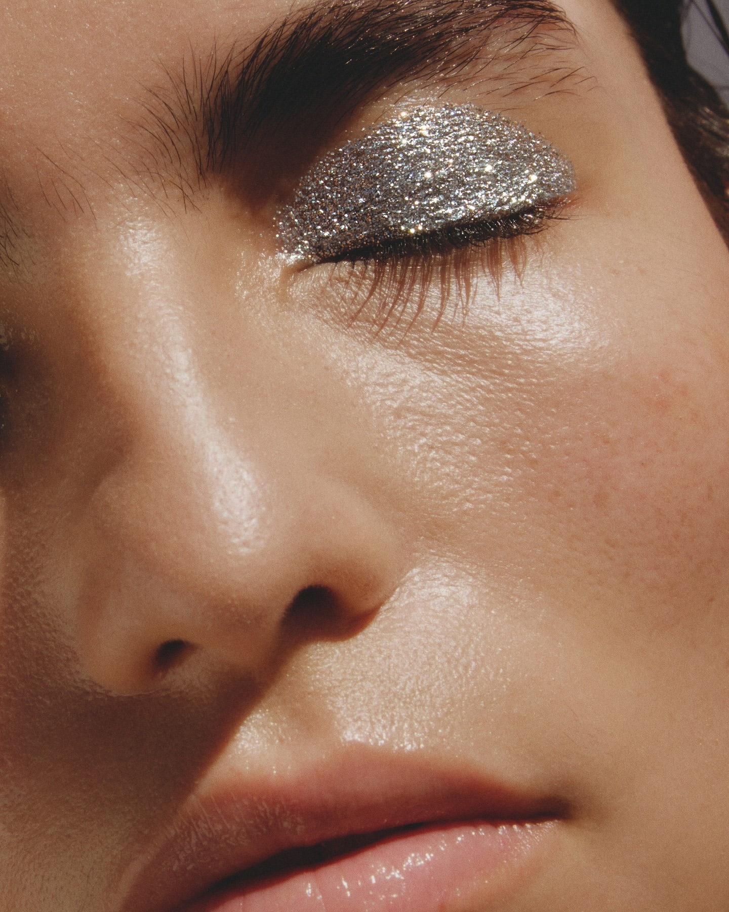 Close-up portrait of a woman with silver glitter eye makeup covering the eyelid, set against a neutral background, showcasing beauty.
