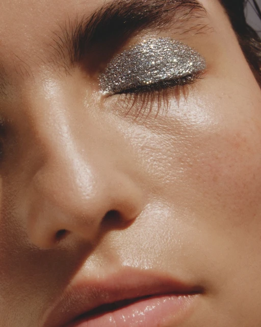 Close-up portrait of a woman with silver glitter eye makeup covering the eyelid, set against a neutral background, showcasing beauty.