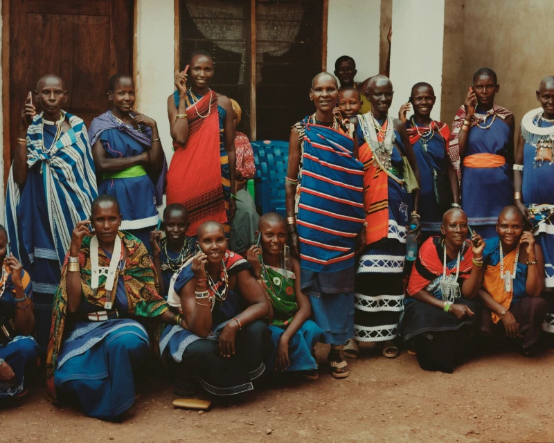 Group of Maasai women, some holding cell phones, pose outdoors in traditional colourful clothing and beaded jewelry.