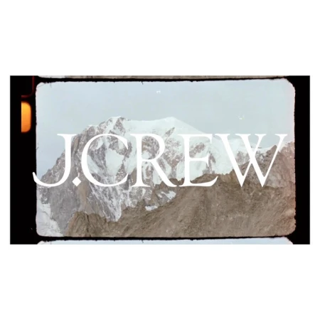 Snow-covered mountain range with a dramatic peak, set against a pale sky, creating a cool and scenic outdoor vista. J.Crew logo.