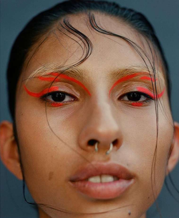Close-up portrait of a young adult female with red eyeliner and a nose ring against a gray backdrop in a beauty-style photograph.