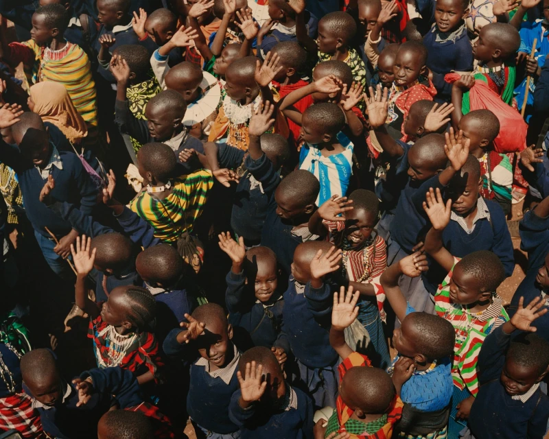 Large group of African children with their hands raised, wearing various patterned tops and blue bottoms, shot from a high angle.